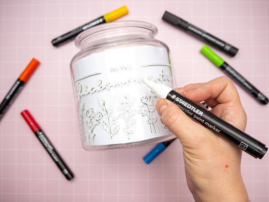 Jemand überträgt mit einem weißen Paintmarker von Staedtler ein Lettering von einer Vorlage auf ein Bonbonglas. Drumherum liegen weitere, farbige Paintmarker. Der Untergrund ist ein rosafarben mit weißem Karomuster. Foto: Johanna Rundel 