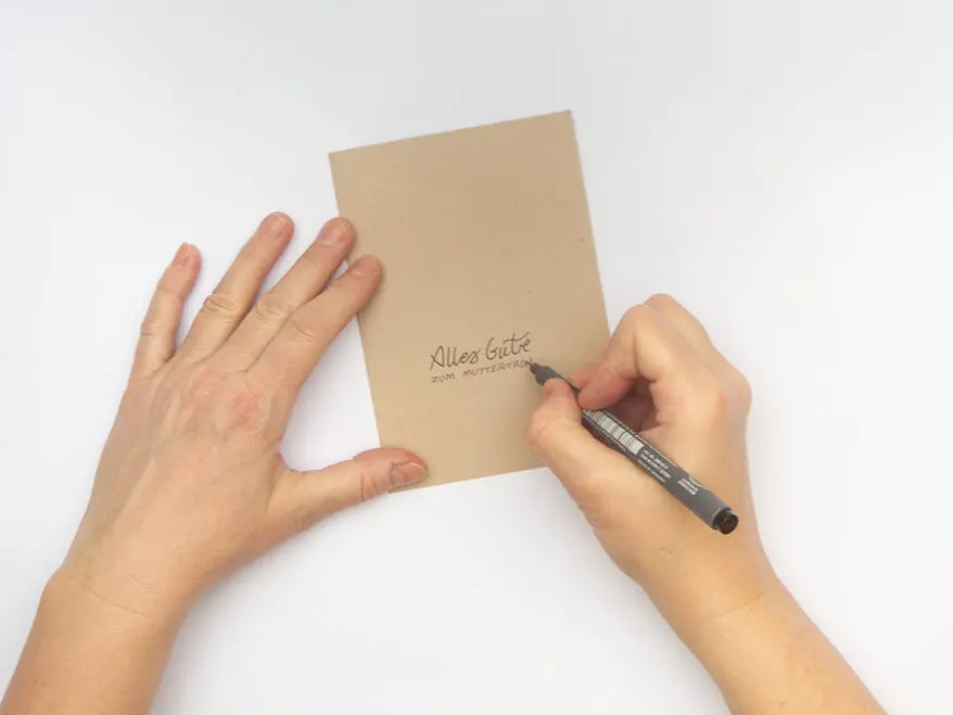 Jemand lettert den Schriftzug "Alles Gute zum Muttertag" auf eine Klappkarte aus Kraftkarton von Artoz. Der Untergrund ist weiß. Foto: Johanna Rundel