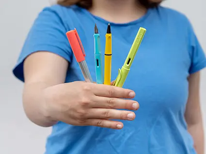 Ein Teenager-Mädchen im blauen T-Shirt hält eine Auswahl bunter Füller und Tintenroller von Lamy in die Kamera.