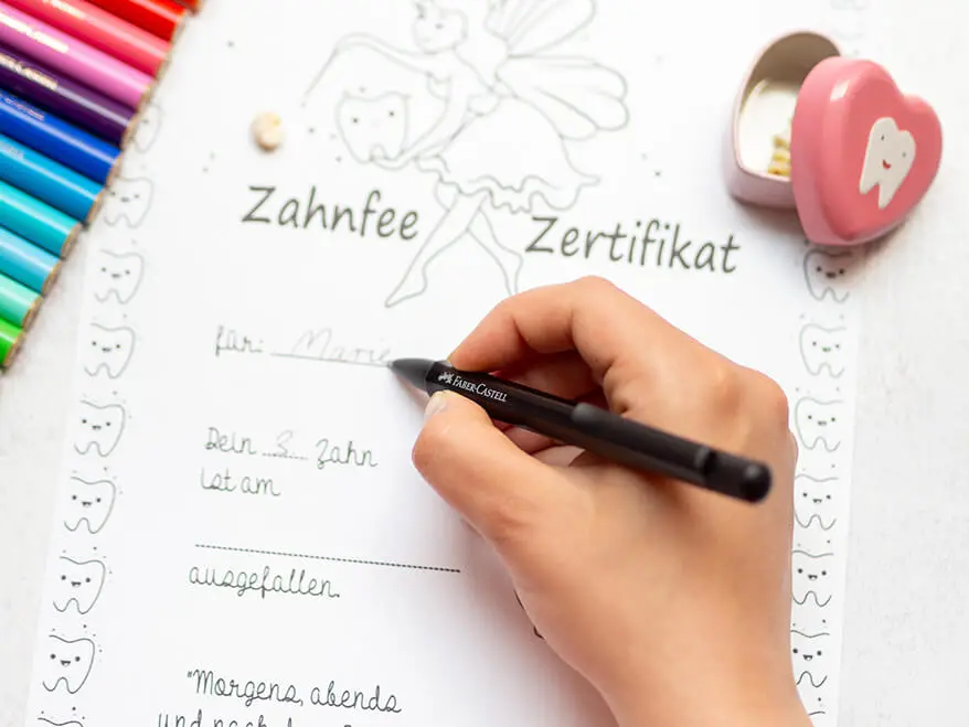 Jemand füllt mit einem Kugelschreiber von Faber-Castell ein Zahnfee-Zertifikat aus. Außerdem zu sehen: eine rosa Zahndose und bunte Filzstifte. Foto: Johanna Rundel