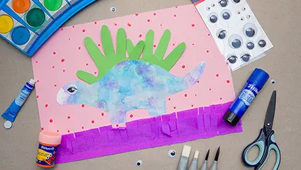 Auf einem grauen Untergrund liegt ein, aus einem Handabdruck selbstgestaltetes, Dinosaurier-Bild. Drumherum sind verschiedene Bastelmaterialien und Farbstifte von Pelikan und Herlitz verteilt. Foto: Johanna Rundel