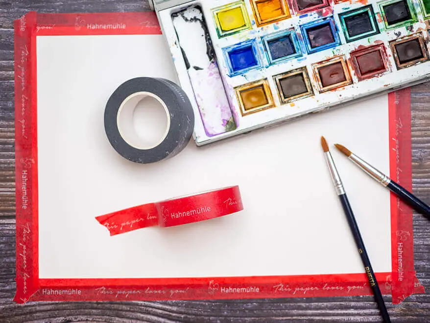 Auf einem dunklen Holztisch ist ein Aquarellpapier von Hahnemühle mit rotem Washitape aufgezogen. Pinsel und Aquarellfarbkasten liegen daneben. Foto: Johanna Rundel 