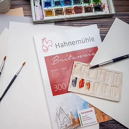 Auf einem dunklen Holztisch sind verschiedene Aquarellpapiere von Hahnemühle ausgebreitet. Pinsel und Aquarellfarbkasten liegen daneben. Foto: Johanna Rundel 