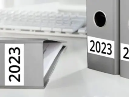Drei graue Aktenordner mit der Jahreszahl 2023 auf dem Rücken stehen auf einem hellen Schreibtisch. Im Hintergrund ist eine weiße Tastatur zu sehen.