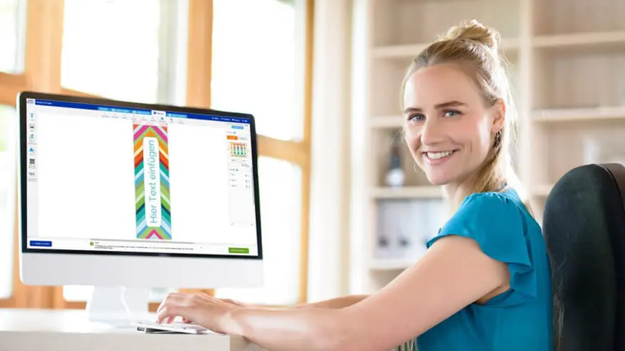 Eine junge, blonde Frau mit blauem T-Shirt sitzt vor Ihrem Computer und gestaltet Etiketten mit der Software von Avery Zweckform.