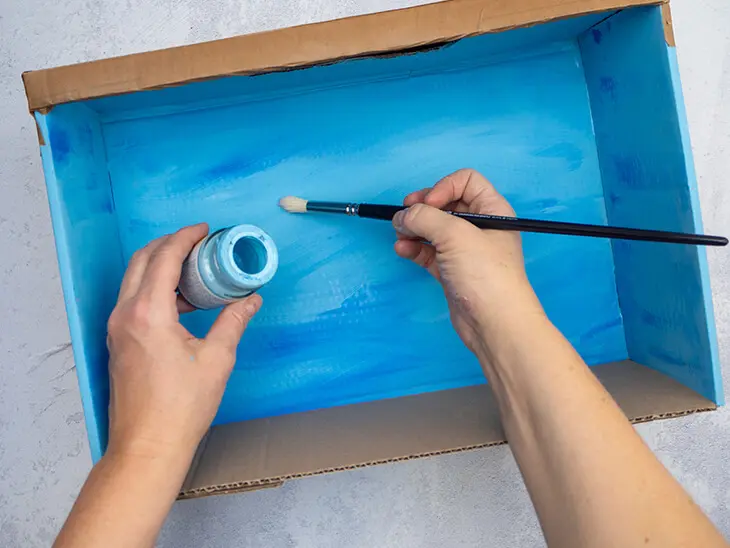 Jemand malt mit einem Pinsel die Innenseite eines Schuhkartons blau an. Foto: Johanna Rundel