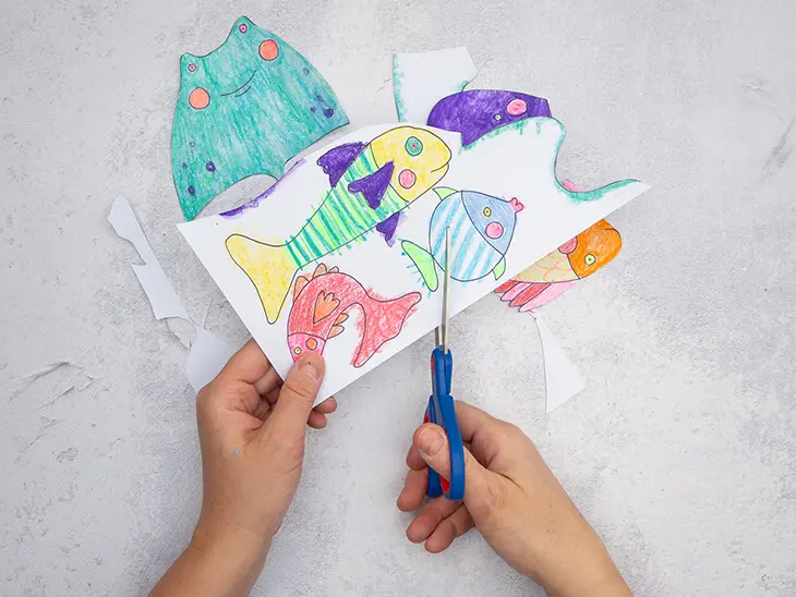 Jemand schneidet mit einer Schere aus einer Vorlage verschiedene mit Bunt- und Wachsmalstiften von STAEDTLER bemalte Fische aus. Foto: Johanna Rundel