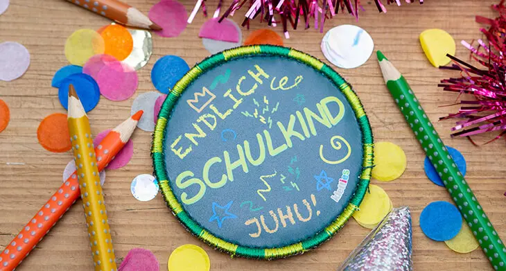 Ein grünes Klettie von ergobag mit der Aufschrift „Endlich Schulkind – Juhu!“ liegt auf einem Holztisch, umgeben von buntem Konfetti. Foto: Johanna Rundel