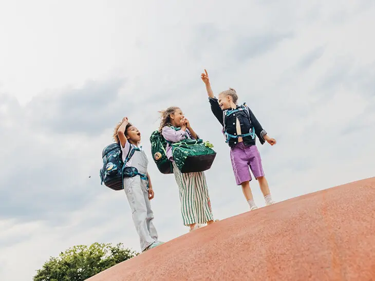 Drei Mädchen im Grundschulalter mit Schulranzen von ergobag auf dem Rücken stehen auf einer Erhöhung und zeigen in den Himmel.
