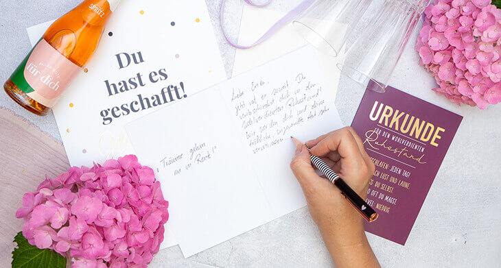Jemand schreibt in eine Grußkarte zum Ruhestand von Grafik Werkstatt einen Text. Weitere Grußkarten und Dekomaterial liegen auf dem grauen Tisch darum verteilt. Foto: Johanna Rundel