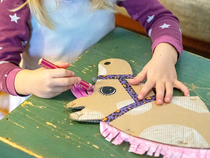 Ein Mädchen hockt vor einer grünen Kiste und malt mit einem rosa Buntstift von STAEDTLER ein selbstgebasteltes Steckenpferd aus Pappe an. Foto: Johanna Rundel)