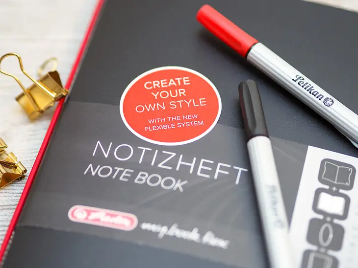 Ein schwarzes my.book flex von herlitz mit einem roten Aufkleber auf dem „Create your own Style” steht. Daneben liegen zwei Fineliner von Pelikan. Foto: Johanna Rundel