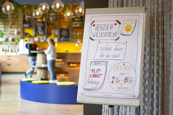 Ein Flipchart mit dem Text „Herzlich Willkommen“ steht im Vordergrund, im Hintergrund sieht man zwei Menschen an einem Stehtisch.)