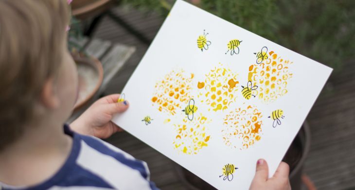 Ein Kind hält ein Blatt Papier in der Hand, auf das Bienenwaben und Bienen gemalt sind.)