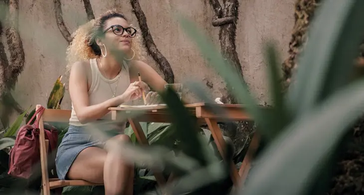 Blick durch Pflanzenblätter auf eine junge Frau, die mit einem Notizbuch von LEUCHTTURM1917 an einem Holztisch sitzt und nachdenkt.