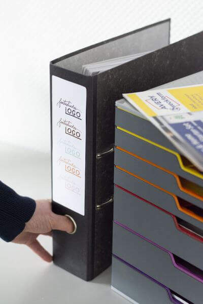 Neben einer Schubladenbox steht ein beschrifteter, schwarzer Ordner. Auf der Schubladenbox liegen Etiketten der Marke Avery Zweckform. Ein Arm ragt ins Bild und greift nach dem Ordner.