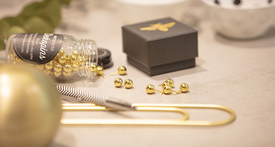 Auf einem Tiusch liegt ein umgekipptes Glas mit goldenen Perlen, welche aus dem Glas herausgefallen sind. Daneben steht eine kleine Schachtel, es liegt ein Kugelschreiber auf dem Tisch, eine große goldene Büroklammer und eine golden Weihnachtskugel. 