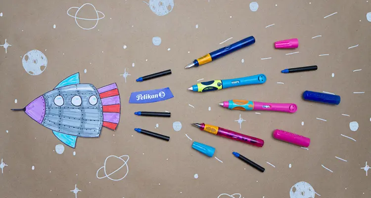 Mehrere Schreiblernstifte von Pelikan liegen auf einer Weltall-Zeichnung.