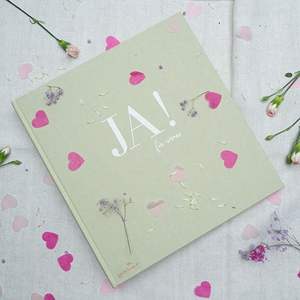 Auf einem hellen Untergrund liegt ein hellgrünes Leinen-Hochzeitsgästebuch von Goldbuch. Darauf ist mit weißer Farbe der Schriftzug „Ja! Für immer“ geprägt. Drumherum liegen pinke und rosa Herzen aus Papier und verschiedene Blumen verteilt. Foto: Johanna Rundel