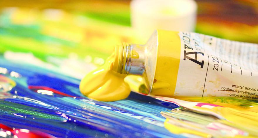 Nahaufnahme einer Tube gelber Acrylfarbe, die offen auf einem bunt bemalten Papier liegt und aus der gelbe Farbe herausläuft.