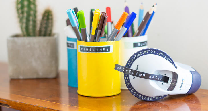 Auf einem Holzregal stehen selbstgemachte Upcycling-Stiftebecher aus alten Chipsdosen in den Farben gelb, petrol und grau. In den Stiftebechern stehen bunte Fineliner und Buntstifte. Im Vordergrund liegt ein Prägegerät von Dymo, aus dem ein Stück Prägeband ragt, auf dem „Upcycling rockt“ seht. Im Hintergrund sieht man verschwommen einen Kaktus.