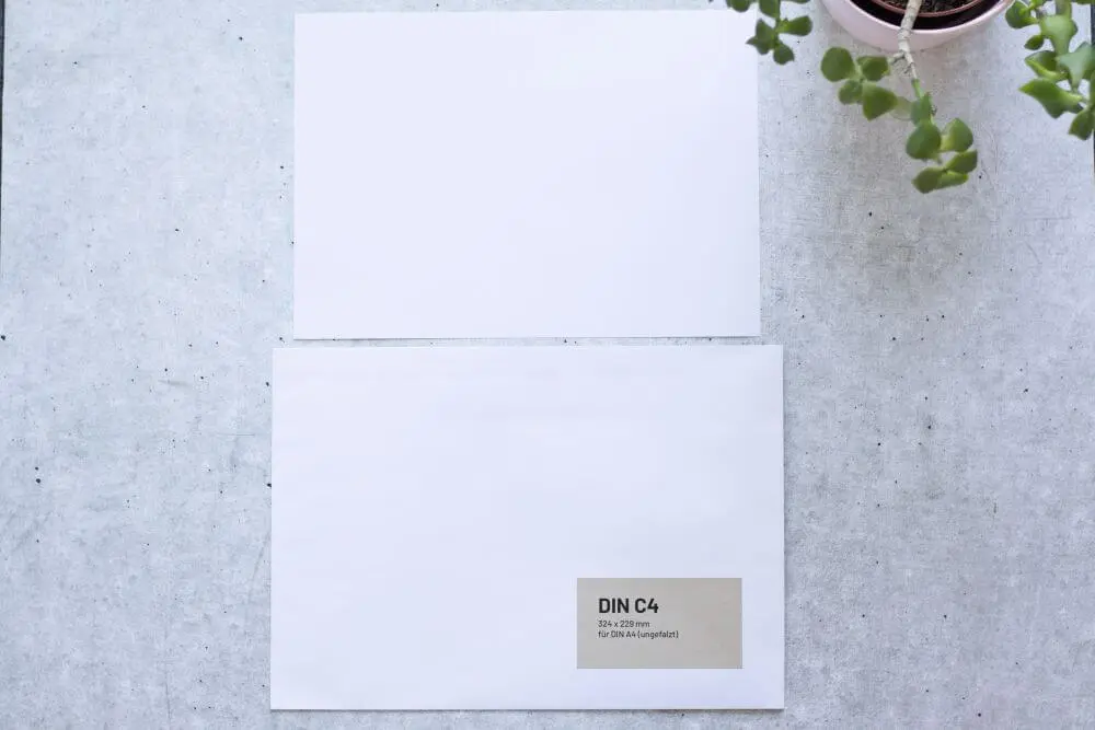 Auf grauem Grund liegt ein DIN A4-Papier und eine dazu passende Versandtasche DIN C4. Auf der Versandtasche ist ein Aufkleber aufgebracht mit folgenden Infos: DIN C4, 324x229 Millimeter, für DIN A4 (ungefalzt). Rechts oben ragt der Teil einer Pflanze ins Bild.