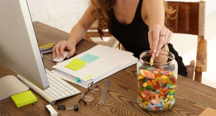 Frau greift in Glas mit bunten Süßigkeiten auf ihrem Schreibtisch.