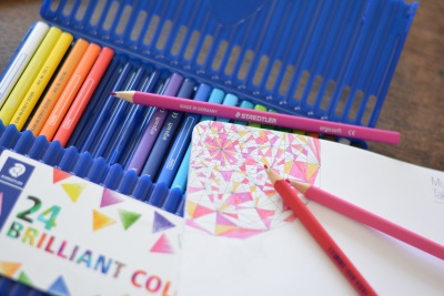 Auf einer aufgeklappten Packung Buntstiften der Marke Staedtler liegt ein Block, auf dem bunte Kästen gemalt sind.