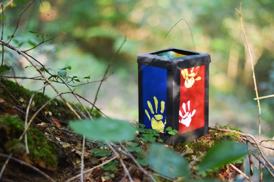 Auf einem Waldboden steht eine gebastelte Kinderlaterne. Eine Seite ist mit blauem, eine andere mit roten Transparentpapier beklebt, auf denen Kinder-Handabrücke aufgebracht sind.