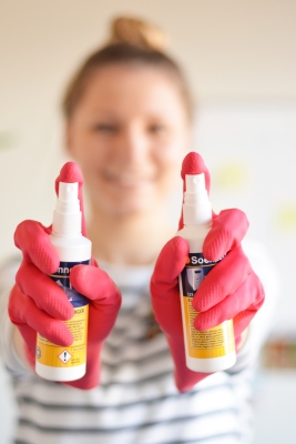 Nahaufnahme von 2 Händen in Gummihandschuhen, die jeweils eine Flasche Desinfektionsmittel in die Kamera halten.