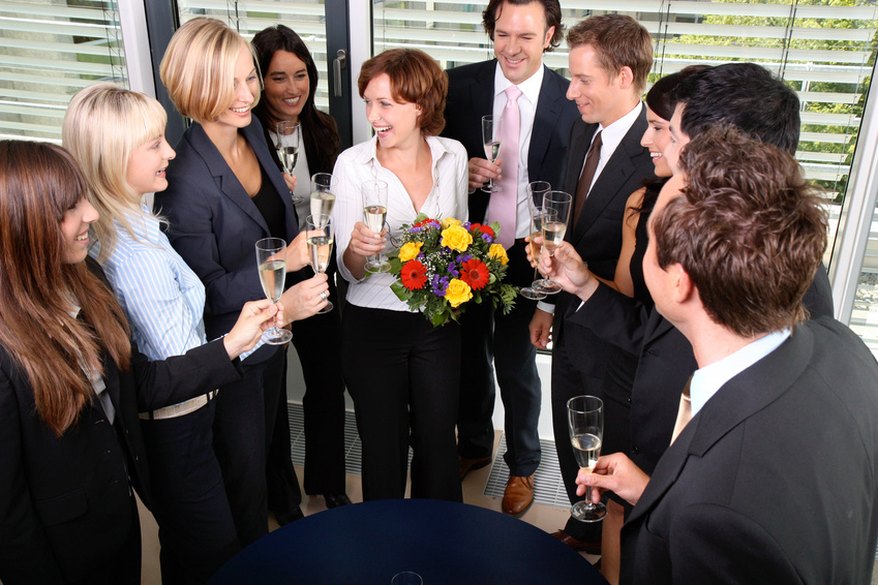 Gruppe im Büro prostet einer Frau mit Blumenstrauß zur Begrüßung zu.