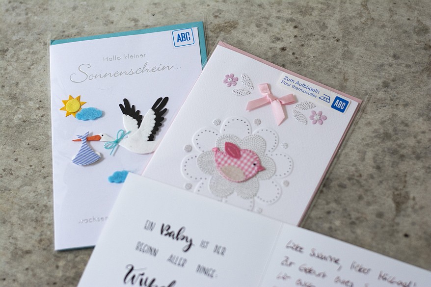 Glückwunschkarten zur Geburt der Marke ABC. Auf einer Karte ist ein Storch abgebildet, auf der anderen Karte ein rosa Vogel.