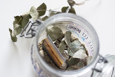 Foto von oben in ein geöffnetes Weckglas, welches mit Sand, Muscheln, Efeu und einem kleine  Foto besfüllt ist. Neben dem Glas liegt eine Efeu-Ranke auf dem Tisch.