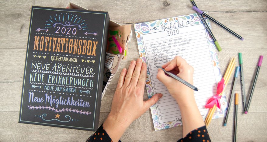 Auf einem Holztisch liegt eine Kiste, die mit Metallic Markern von Stabilo beschriftet wurde. Es handelt sich um eine Motivationsbox. Links davon schreibt eine Person gerade eine Bucket List für das Jahr 2020. Um die Liste liegen verschiedene Marker der Marke Stabilo.