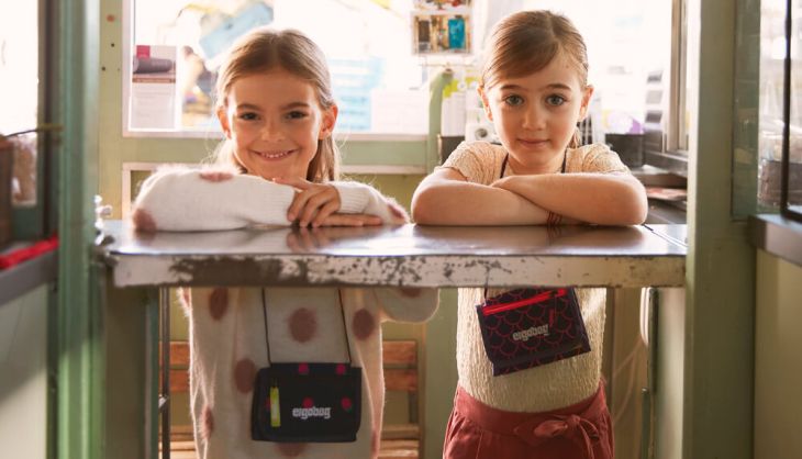 Zwei Mädchen im Grundschul-Alter lächeln in die Kamera. Beide haben einen Brustbeutel der Marke ergobag um.