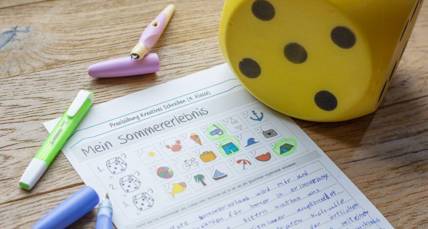 Auf einem Holztisch liegt ein Übungsblatt mit Schreibübungen, daneben liegen ein großer Würfel und 2 Füller.