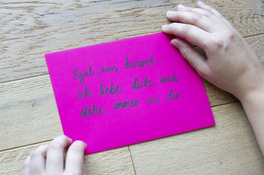 Auf einem Untergrund aus Holz liegt ein rosafarbenes Blatt Papier. Das Blatt wird von zwei Händen gehalten. Darauf ist zu lesen "Egal, was kommt: Ich liebe dich und stehe immer zu dir."