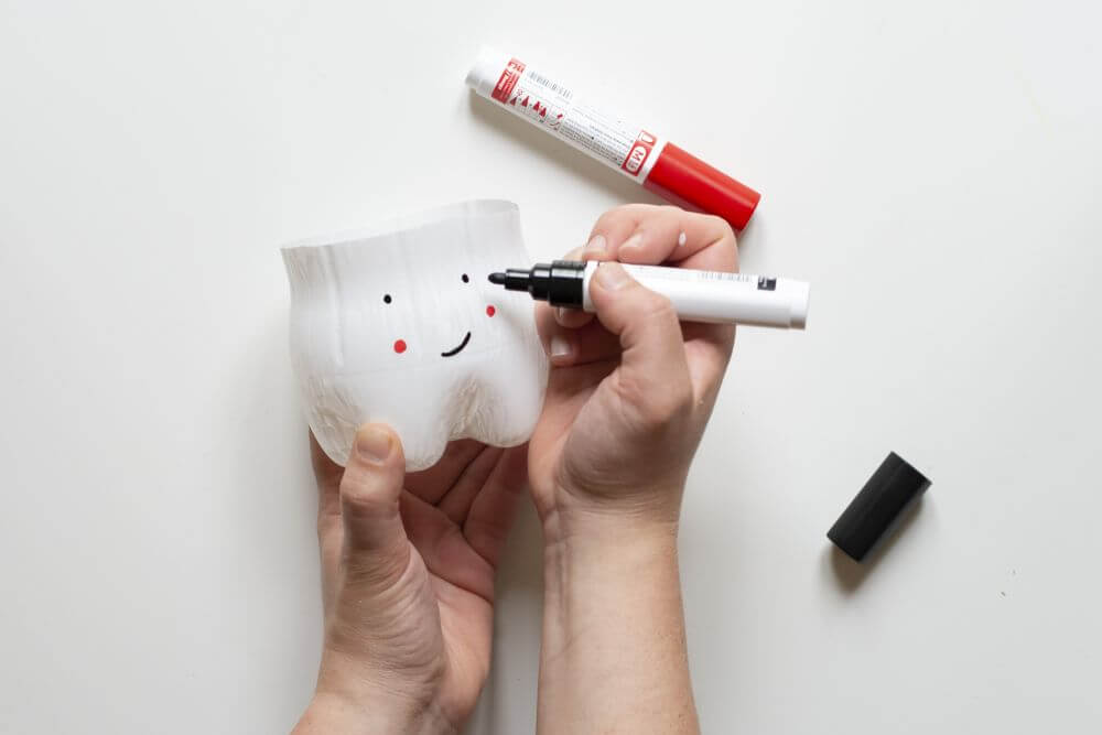 Ein Zahnputzbecher aus einer PET-Flasche wird mit Lackmarkern bemalt. Dieser sieht wie ein Zahn aus.