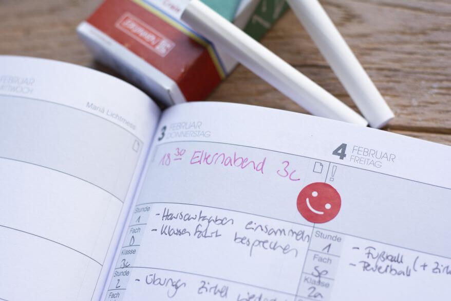 Ein Kalender liegt aufgeschlagen auf einem Holztisch. Darin sind einige Termine notiert. Im oberen Bildrand ist weiße Tafelkreide zu sehen.