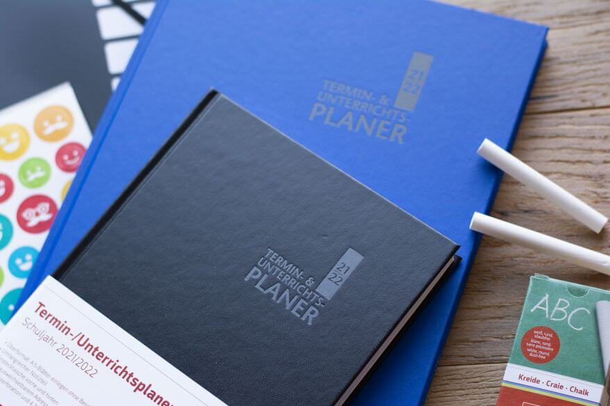 Auf einem Holztisch liegen zwei Termin- & Unterrichtsplaner von Brunnen sowie einige Sticker und Kreide.
