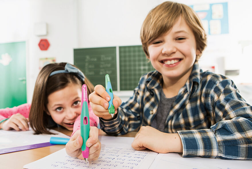 Ein Mädchen und ein Junge sitzen nebeneinander im Klassenzimmer am Tisch. Das Mädchen hält einen Linkshänder Füller von Stabilo nach vorne. Der Junge lacht. Foto: Stabilo