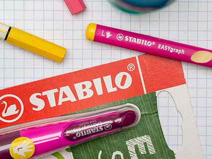 Ein verpackter Linkshänder Füller, ein pinker Linkshänder-Bleistift und ein blauer Linkshänder-Spitzer liegen verstreut auf einem karierten Blatt. Daneben liegen zwei farbige Filzstifte der Marke Stabilo. Foto: Johanna Rundel