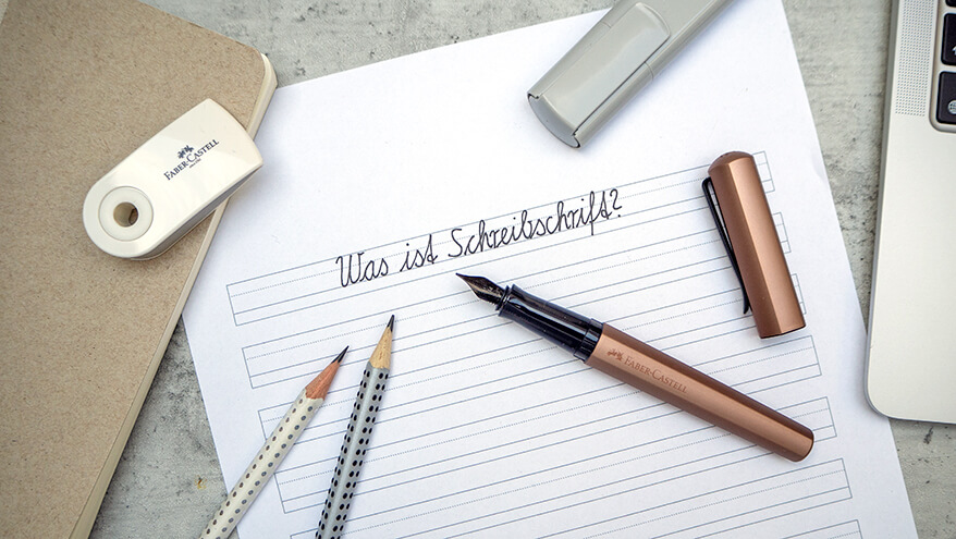 Auf einem Untergrund in Betonoptik sieht man eine Arbeitsplatz-Szenerie. In der Mitte liegt ein Blatt Papier mit der Aufschrift „Was ist Schreibschrift?“, darauf ein kupferfarbener Hexo Füller, zwei Bleistifte und ein grauer Textmarker der Marke Faber-Castell. Rechts im Bild ist ein Laptop zu sehen. Links oben sieht man ein beiges Notizbuch ins Bild ragen, auf dem ein Radiergummi liegt. Foto: Johanna Rundel