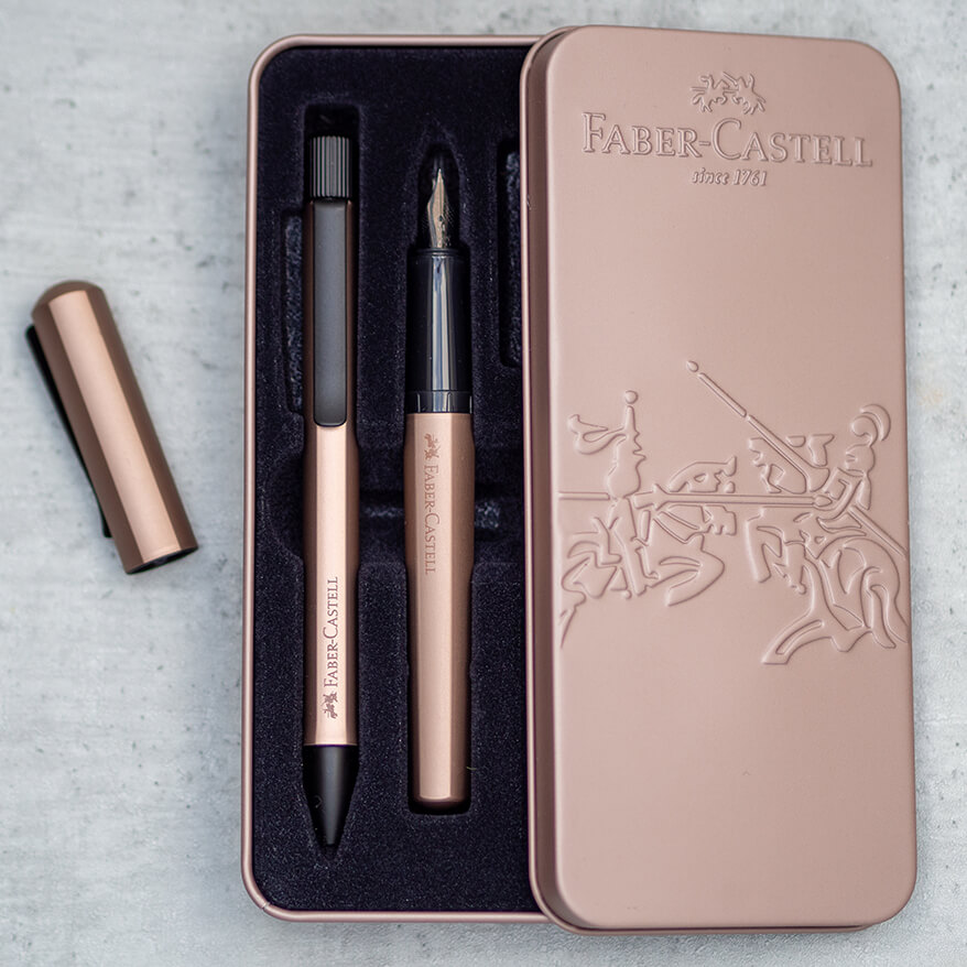 Auf einem Untergrund in Betonoptik liegt eine kupferfarbene Metallschachtel. Der Deckel ist geöffnet und liegt locker seitlich auf. In der Schachtel befinden sich ein ebenfalls kupferfarbener Hexo Füllfederhalter und ein Hexo Ballpen von Faber-Castell. Links neben der Schachtel liegt der Deckel des Füllers. Foto: Johanna Rundel