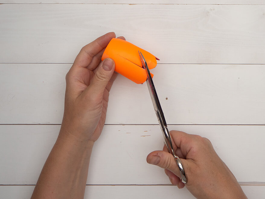 Jemand schneidet eine halbe, orange bemalte kleine Plastikflasche mit einer Schere längs ein und rundet die Streifenenden ab. Der Untergrund ist hell. Foto: Johanna Rundel