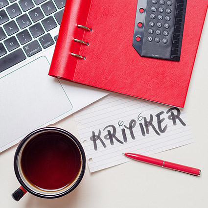 Auf einem weißen Untergrund liegt ein knallrotes Clipbook, dazu ein ausgerissener, linierter Zettel mit dem Wort "Kritiker". Das Buch liegt auf einem aufgeklappten, silbernen Laptop. Auf dem Buch liegt ein flacher Taschenrechner von Filofax. Vor dem Rechner steht eine rote Emaille Tasse mit Tee. Foto: Johanna Rundel