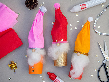 Auf einem grauen Untergrund liegen drei bunte Weihnachtswichtel, die aus Klopapierrollen und Materialien von Baier und Schneider gebastelt wurden. Eine schwarze Schere, mehrere Pearl Maker, Krepppapier und goldener Chenilledraht liegen daneben. Goldene Pailletten sind über den gesamten Untergrund verteilt. Foto: Johanna Rundel