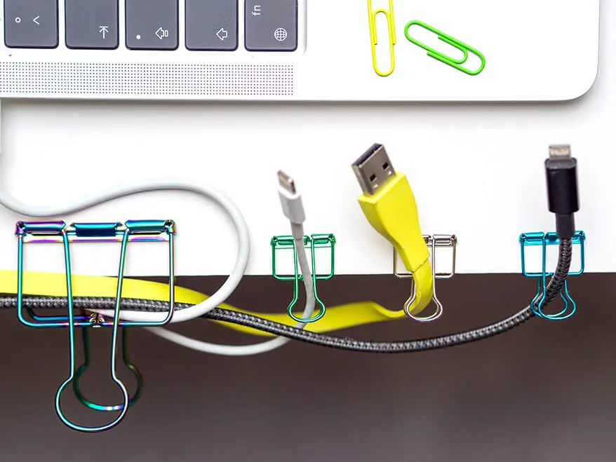 An einer weißen Schreibtischkante klemmen mehrere Kabel und Stecker in bunten Foldback-Klammern von Knorr Prandell. Das Foto ist aus der Vogelperspektive aufgenommen. Im oberen Teil des Bildes ist ein Teil eines silbernen Laptops zu sehen, auf dem zwei bunte Büroklammern liegen. Foto: Johanna Rundel