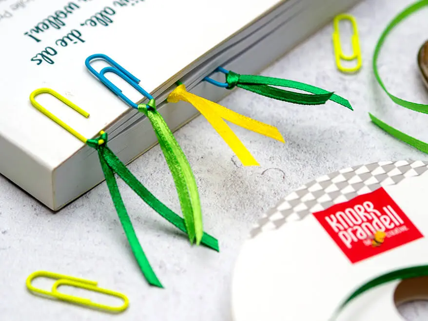 Aus einem Buch ragen mehrere Lesezeichen heraus. Sie wurden aus bunten Büroklammern und dünnem Satinband von Knorr Prandell gebastelt. Eine Rolle des Bandes ist in der rechten unteren Ecke des Bildes zu sehen. Ein paar einzelne Büroklammern sind auf dem grauen Untergrund in Betonoptik verteilt. Foto: Johanna Rundel