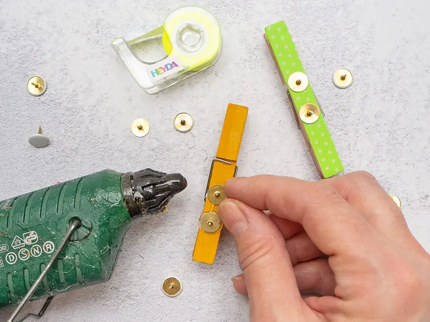Jemand klebt mit Heißkleber eine Reiszwecke auf die Rückseite einer gelb bemalten Wäscheklammer. Links im Bild liegt die grüne Heißklebepistole, rechts eine weitere Klammer, drumherum Washitape-Rollen von Heyda und weitere goldene Reißzwecken. Der Untergrund ist hellgrau. Foto: Johanna Rundel